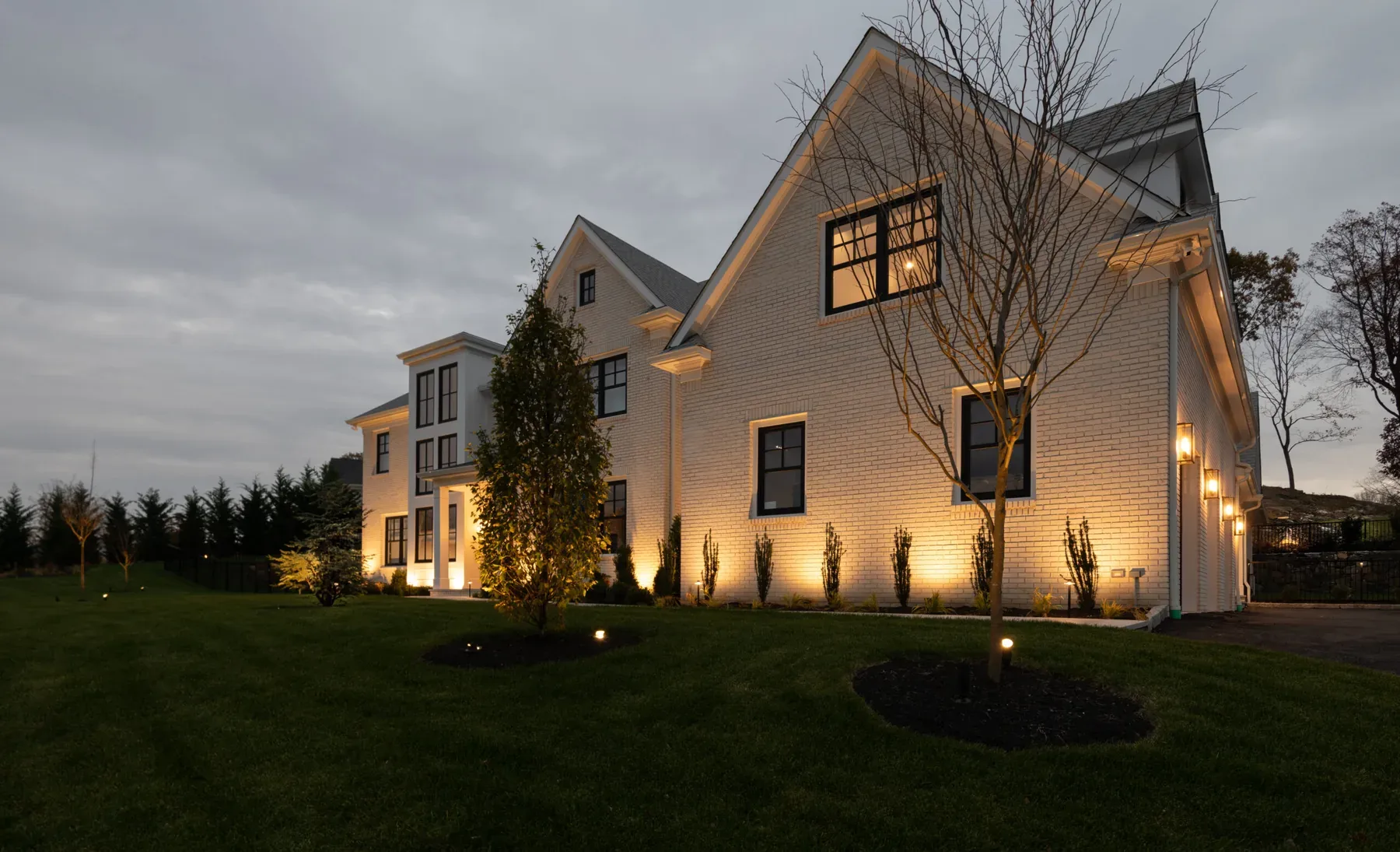 Warm lighting on a gabled house and a young tree beside the lawn.