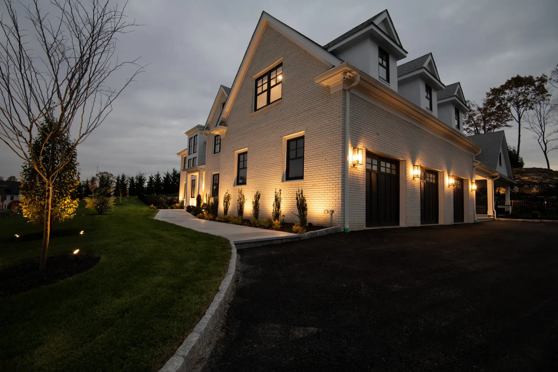 Lit house entrance and driveway bordered by grass and a young tree.