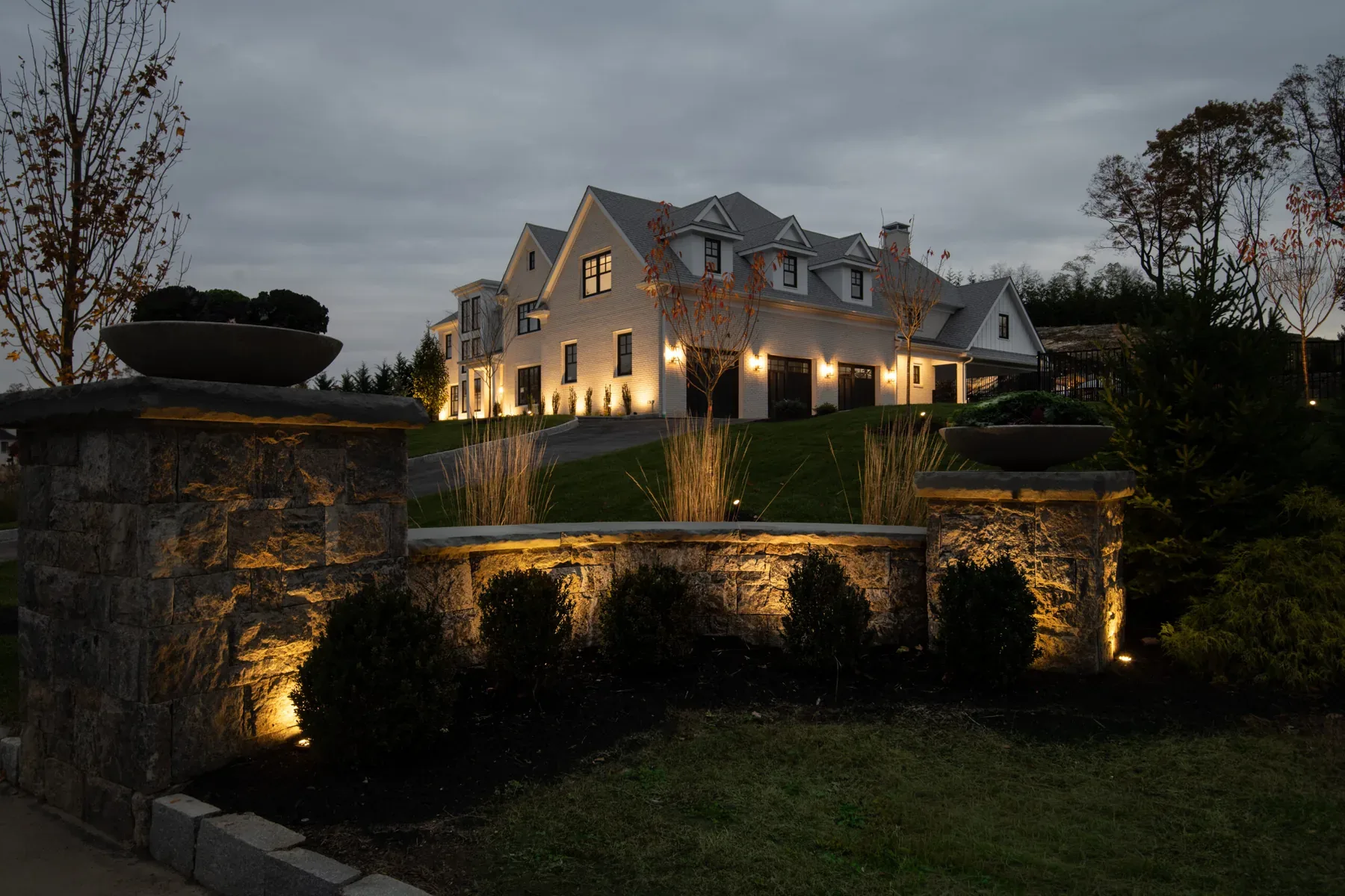 Lit stone entrance wall and planting beds leading toward a house at dusk.