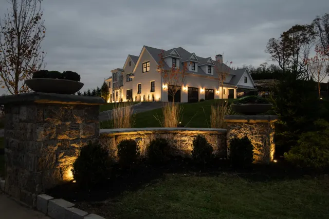 Lit stone entrance wall and planting beds leading toward a house at dusk.