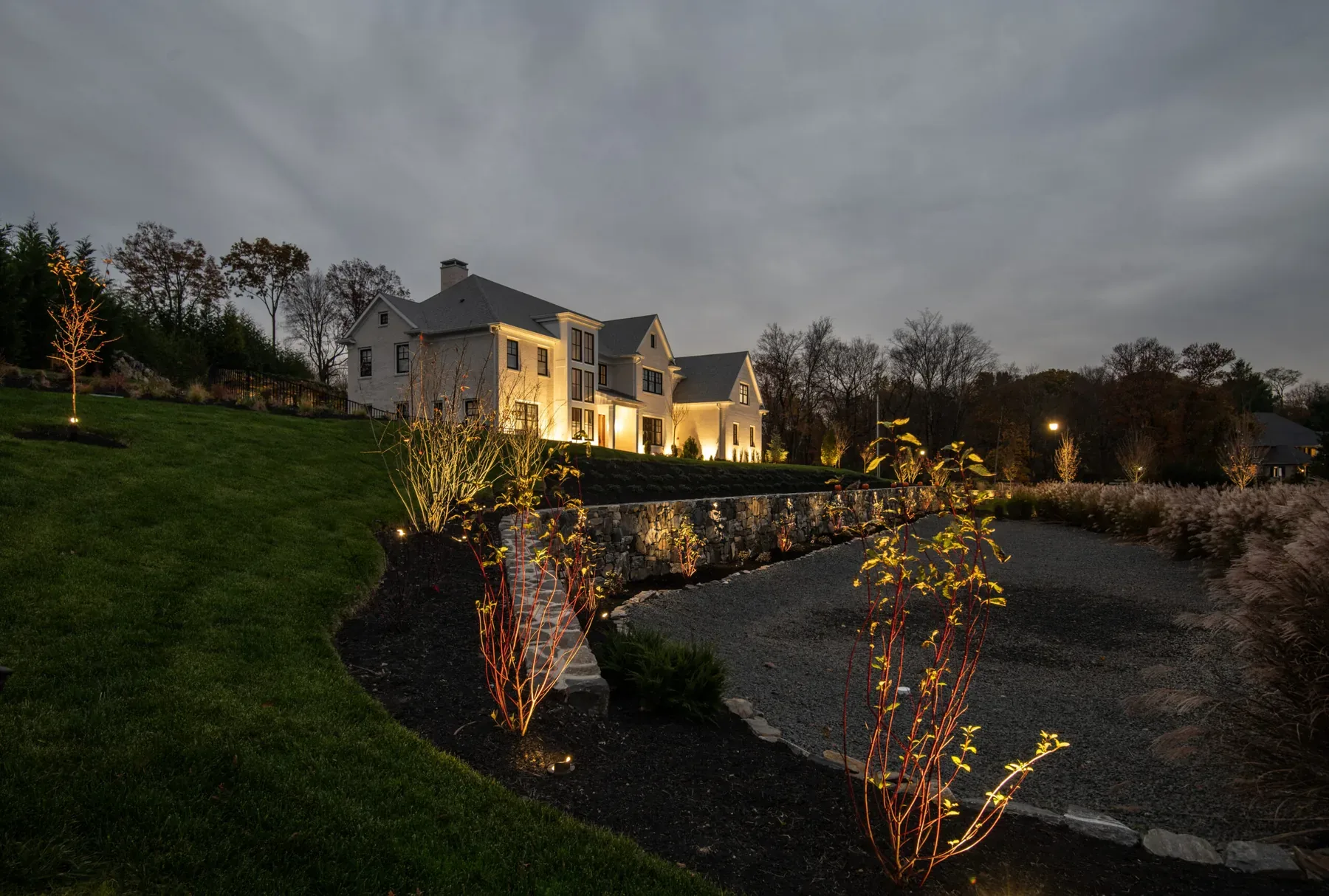 House and young trees illuminated beside a curved stone driveway wall.