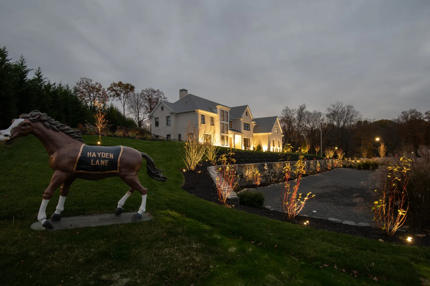 Horse sculpture on a lawn above a lit stone wall and driveway.