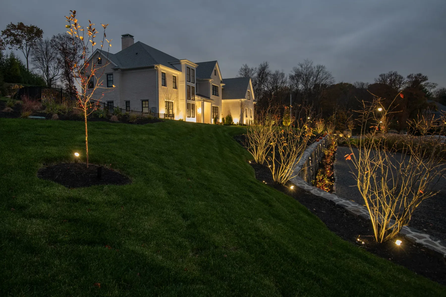 Illuminated house and young trees along a stone wall at dusk.