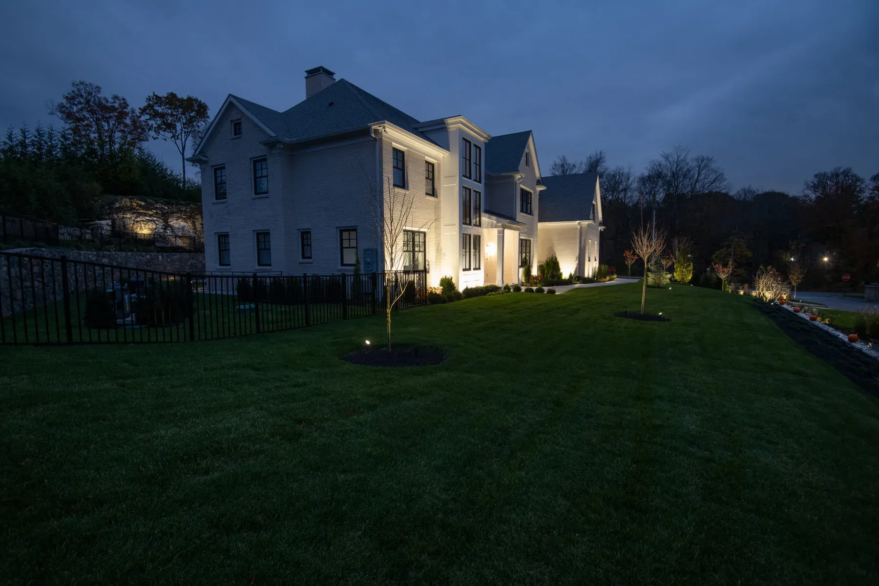 Lit side of a house beside a sloping lawn and black fence at dusk.