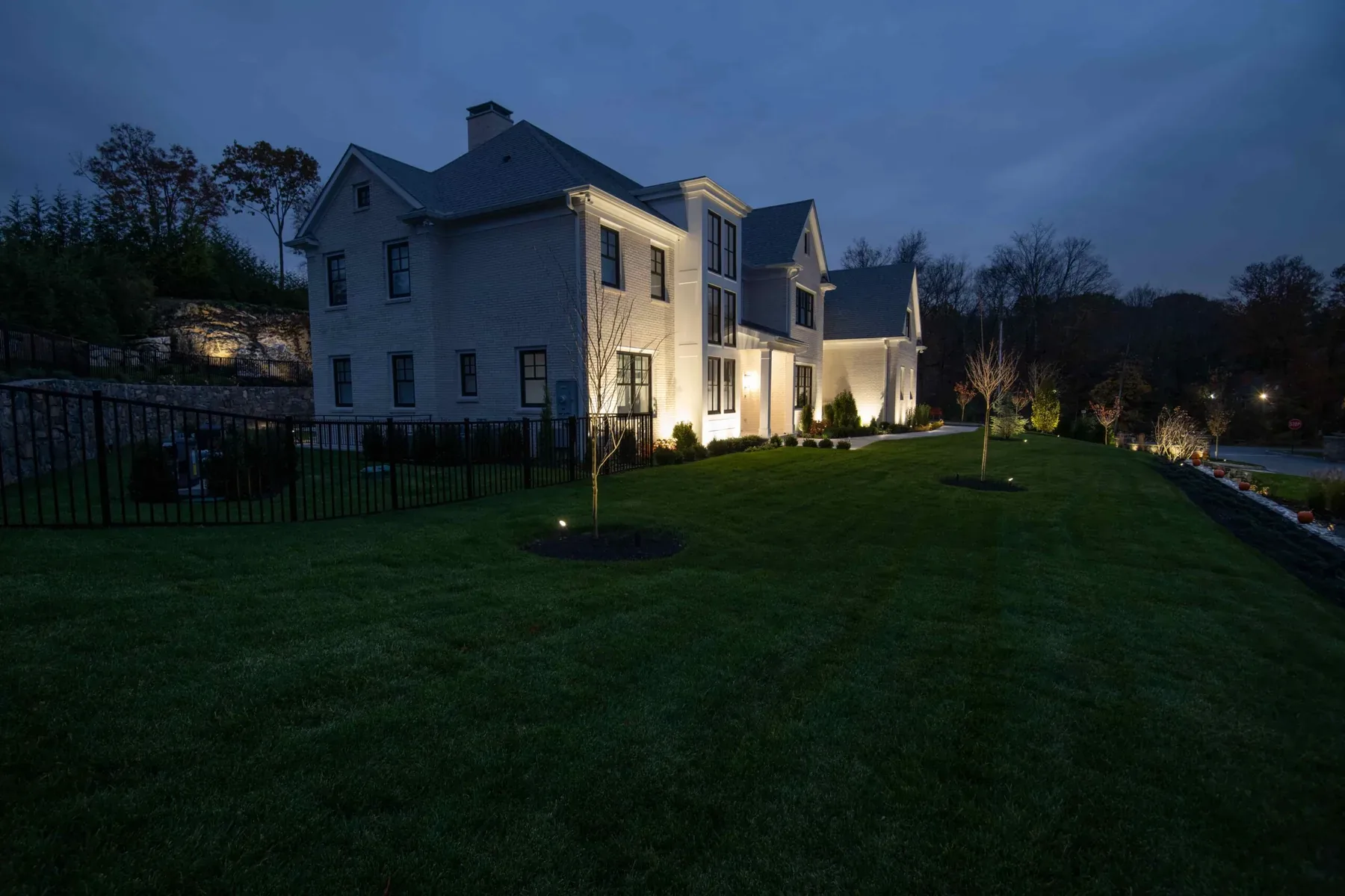 Lit side of a house beside a sloping lawn and black fence at dusk.