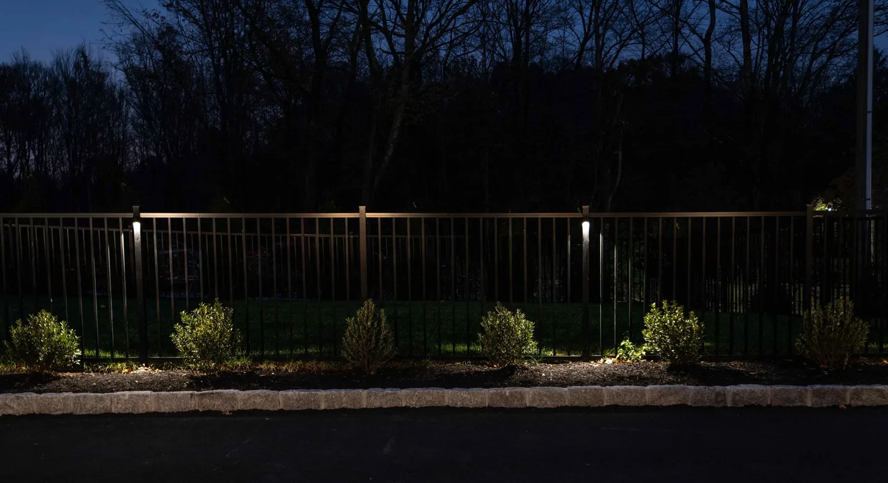Small lights along a black metal fence and planted border after dark.
