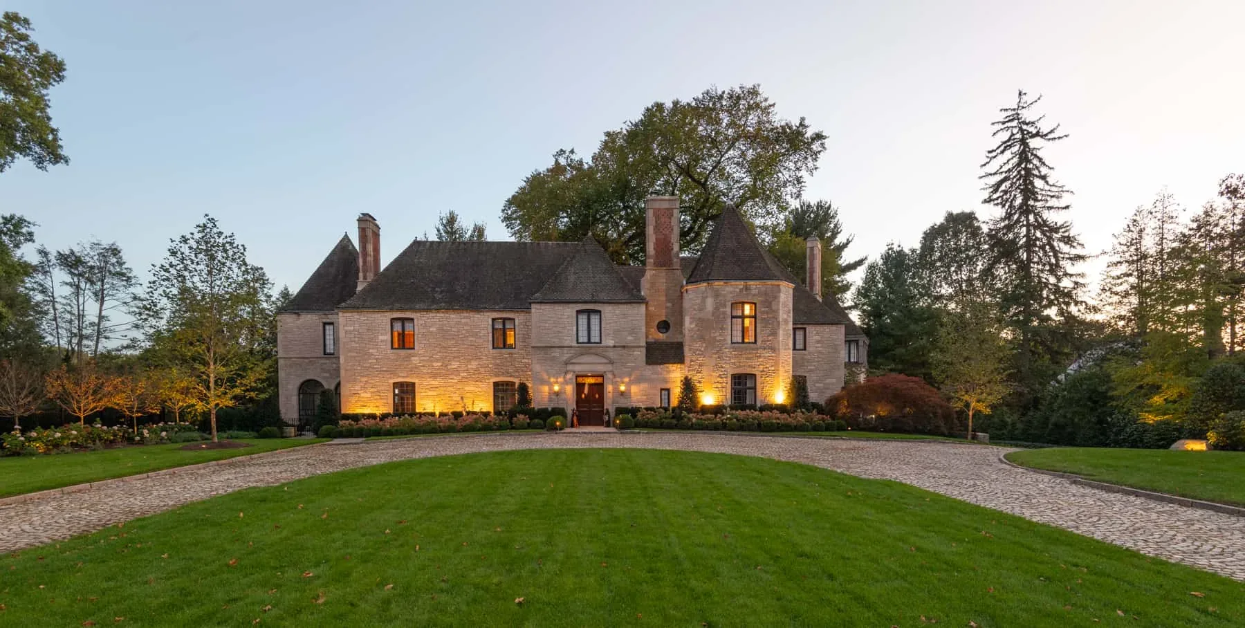 Warm lighting along a stone house facing a broad lawn at dusk.