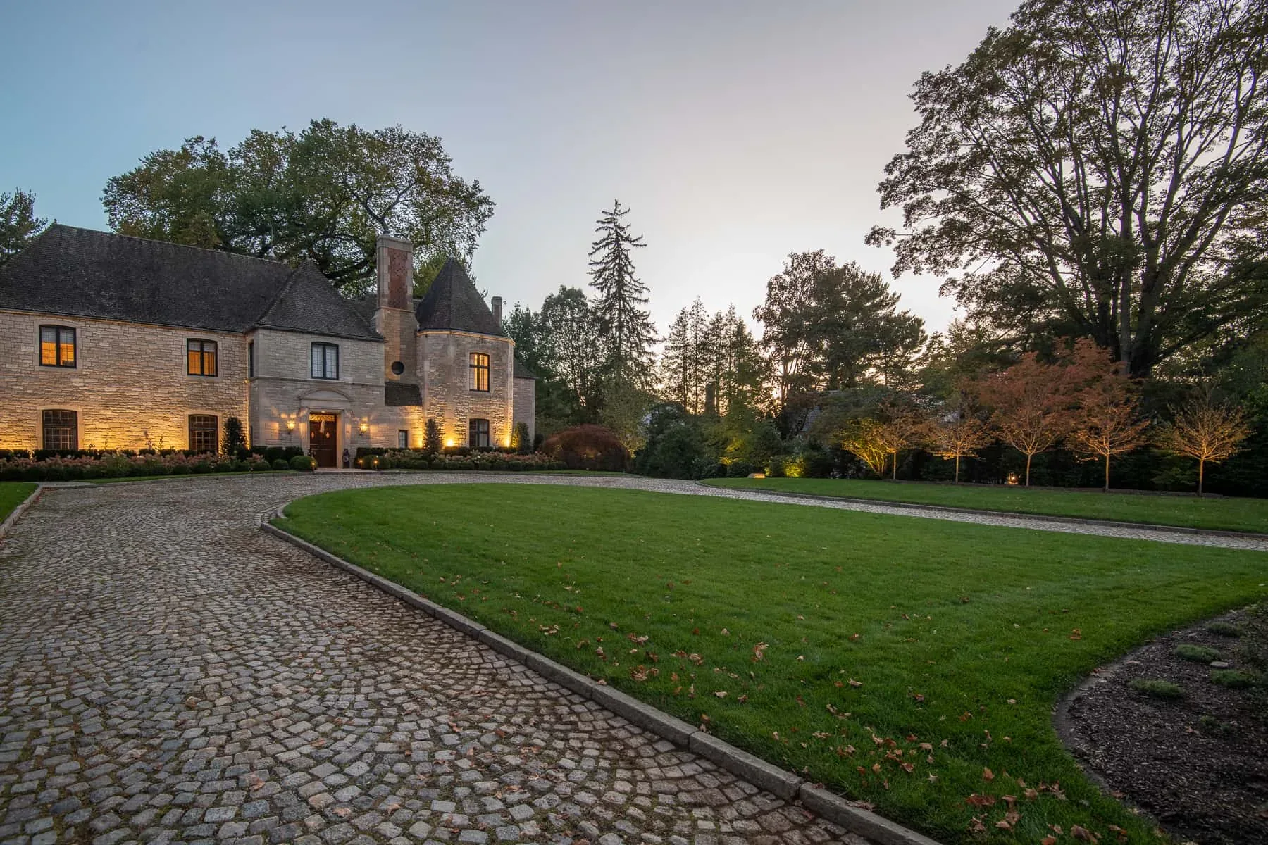 Warm lighting along a stone house facing a broad lawn at dusk.