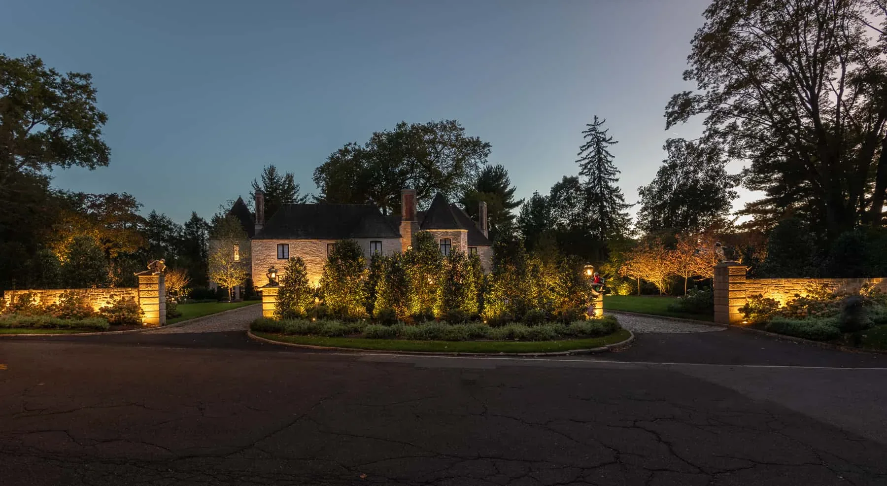 House and landscaped entrance framed by illuminated trees and stone walls.