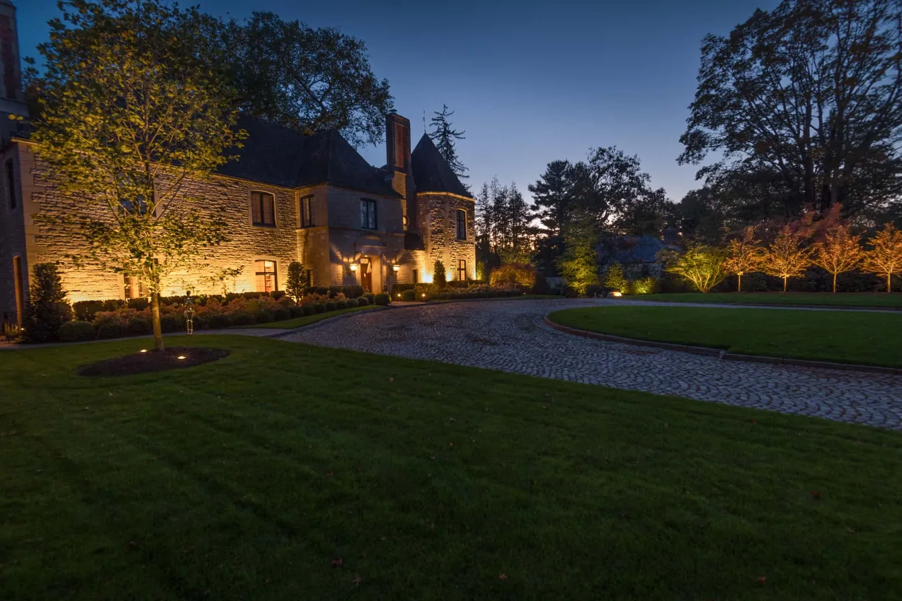 Stone house and trees illuminated beside a curved cobblestone driveway.