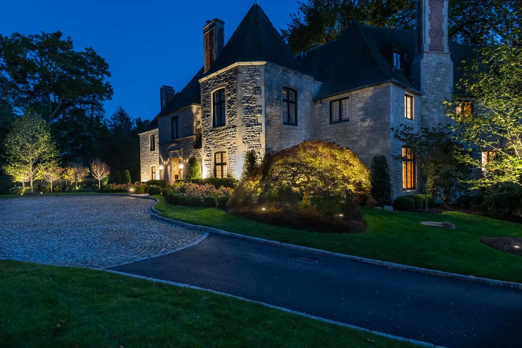 Stone house facade illuminated beside a paved driveway at dusk.
