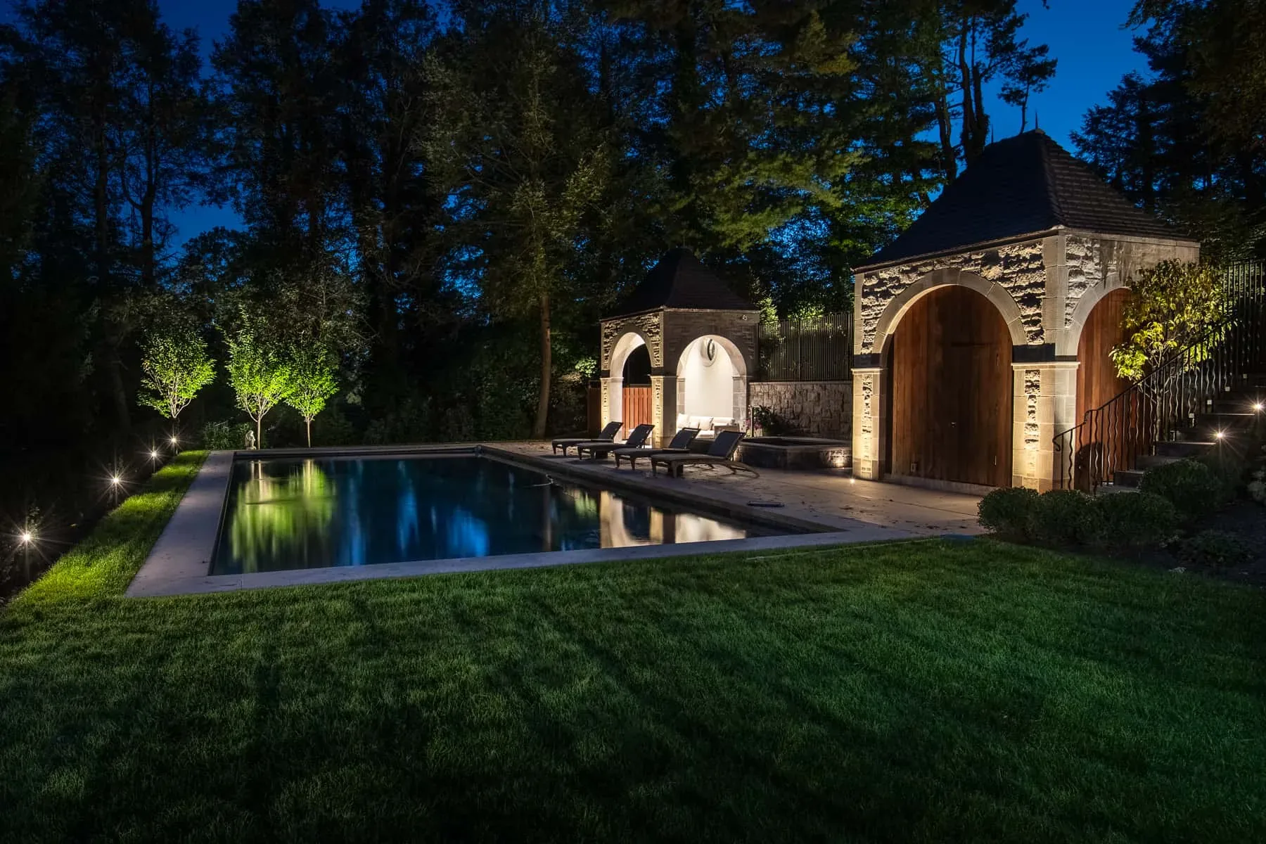 Swimming pool, stone pool house, and lit trees viewed from the lawn at dusk.