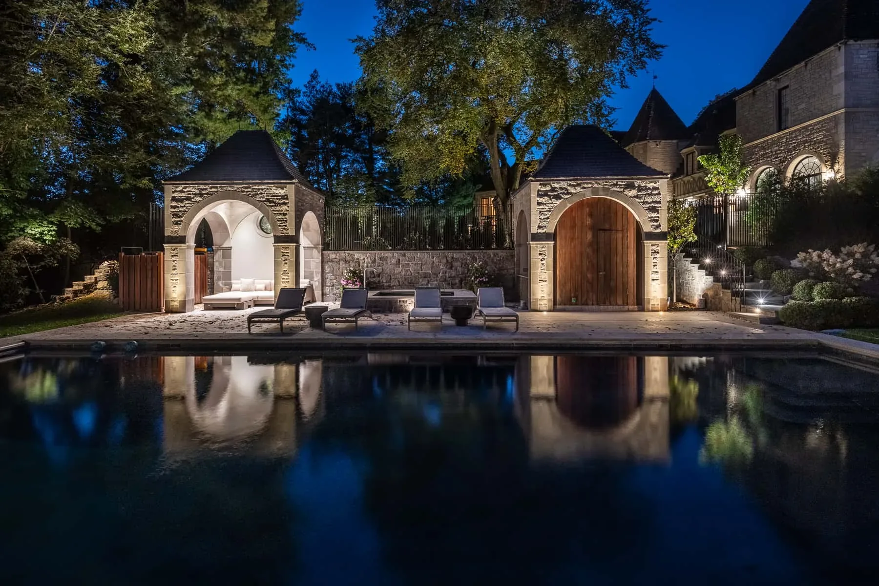 Stone pool house and surrounding trees reflected in a swimming pool at night.