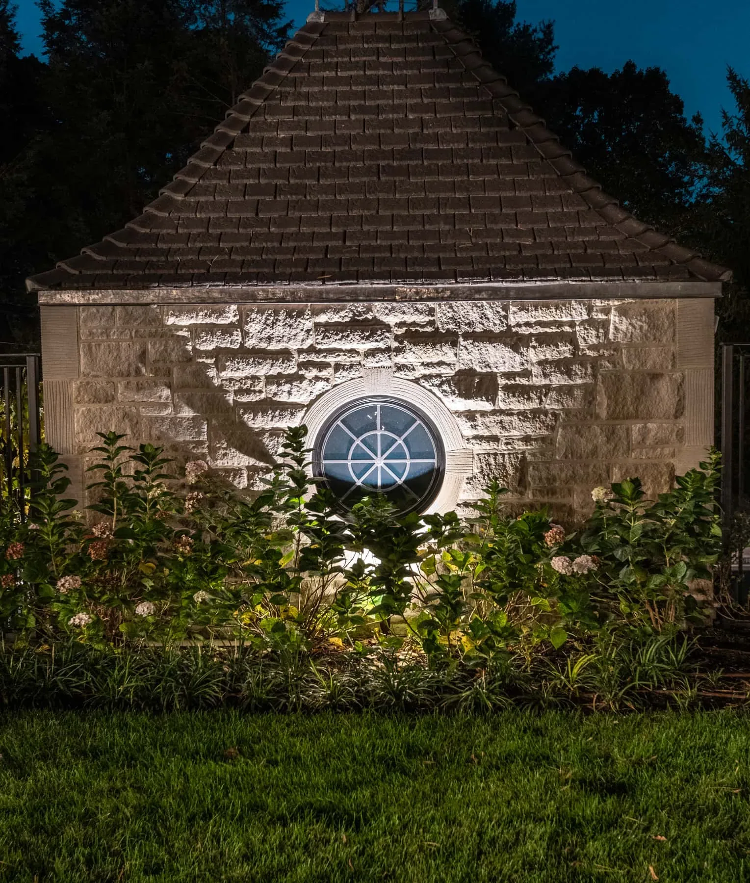 Circular window and stone wall illuminated behind flowering shrubs.
