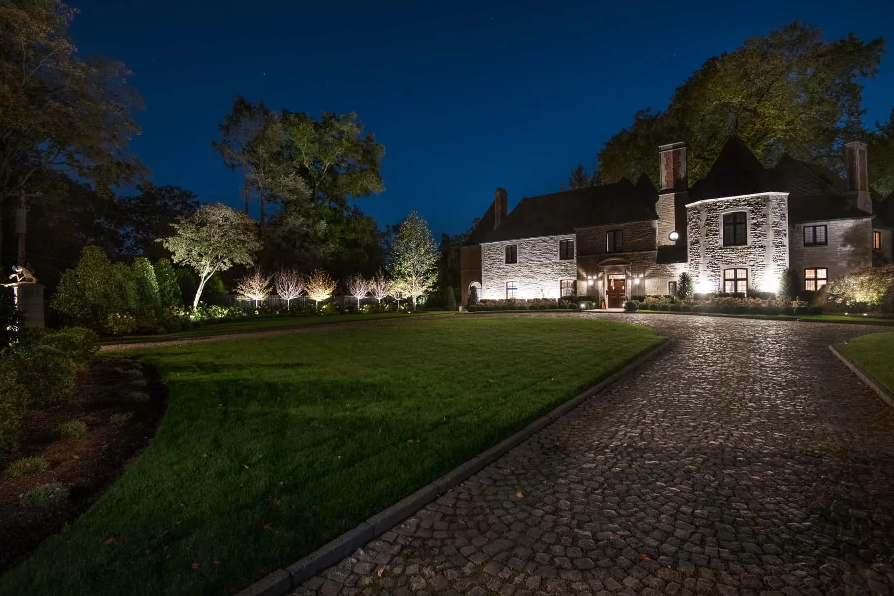 Lit house and trees across a lawn and cobblestone drive at night.