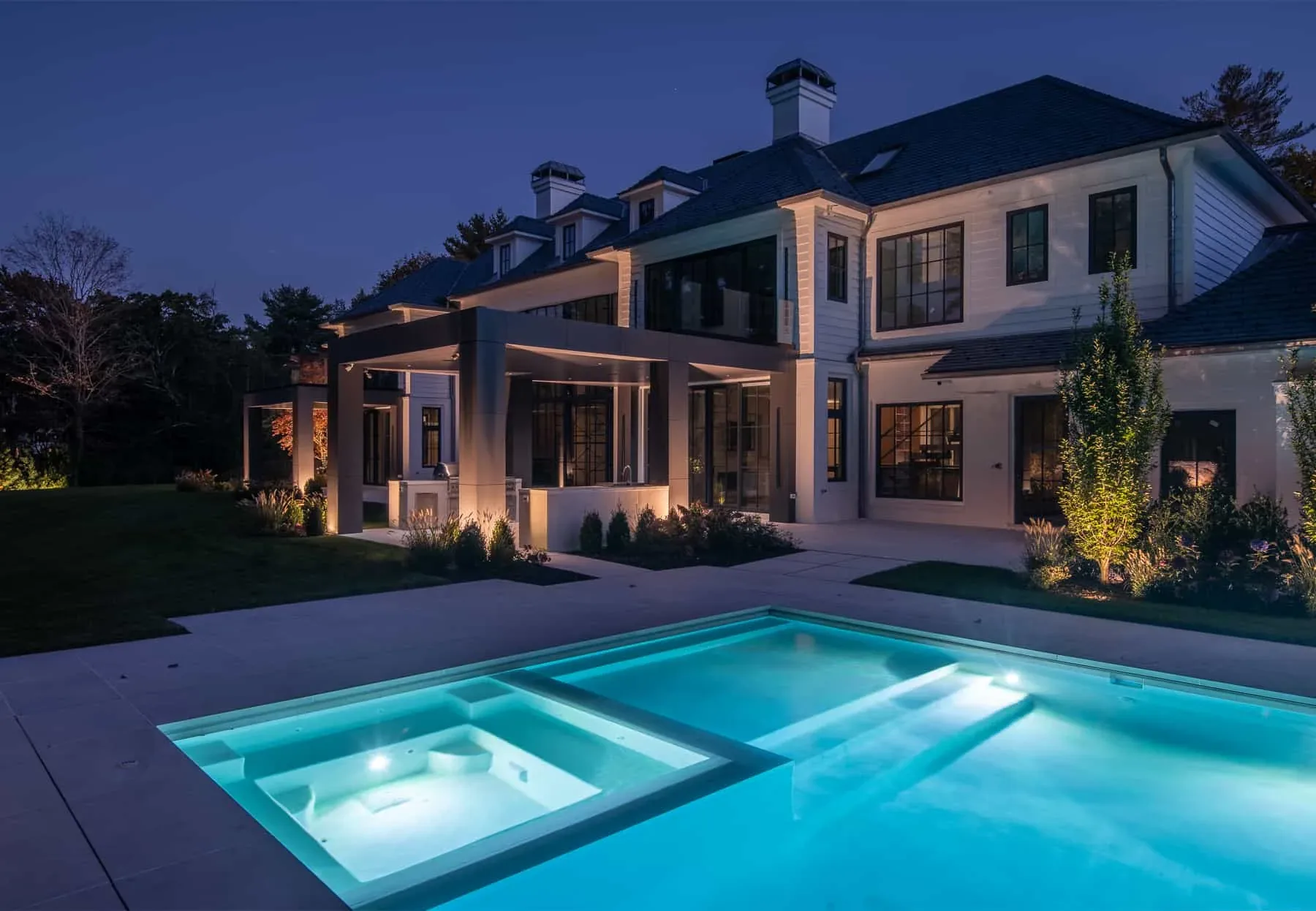 Lit pool and square spa beside the rear terrace of a white house.