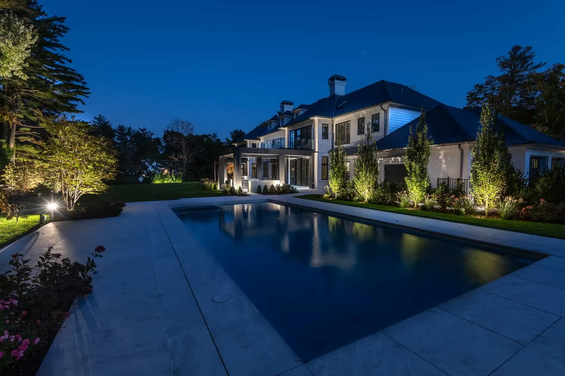 House and illuminated trees reflected in a swimming pool at dusk.