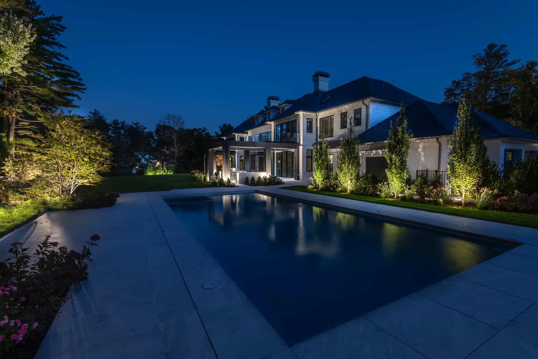 House and illuminated trees reflected in a swimming pool at dusk.
