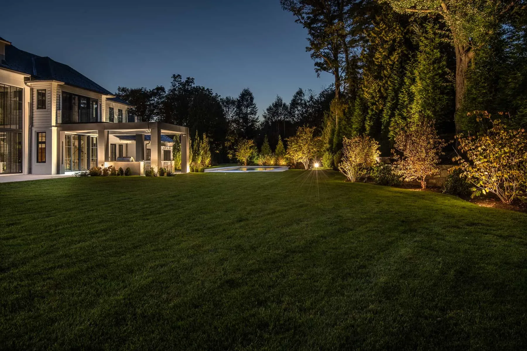 Rear lawn bordered by a lit house, swimming pool, and illuminated trees.