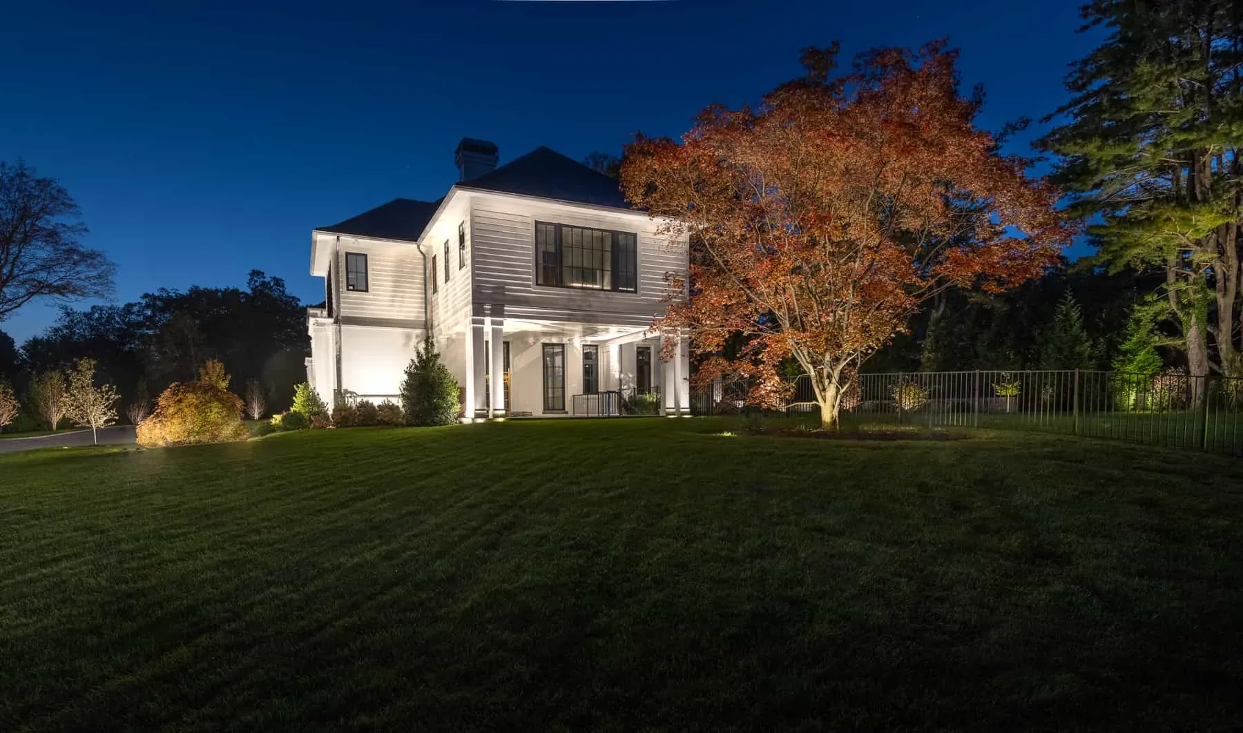 White house and red-leaved tree illuminated beside the lawn at night.