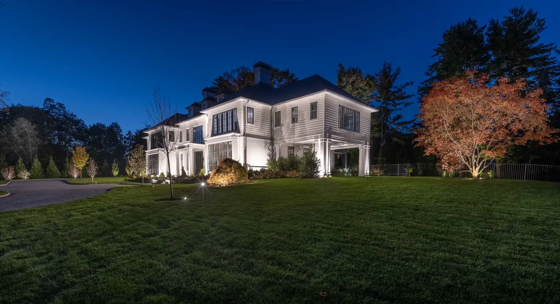 Lighting across a white house facade and trees beside a driveway.