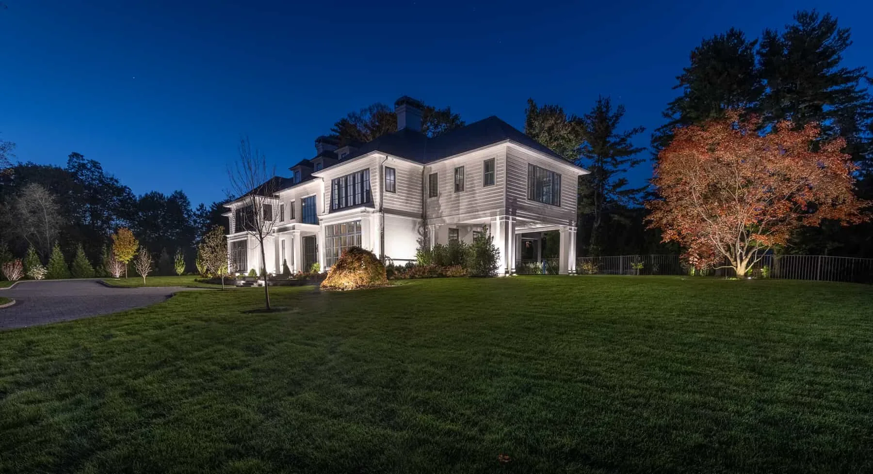 Lighting across a white house facade and trees beside a driveway.