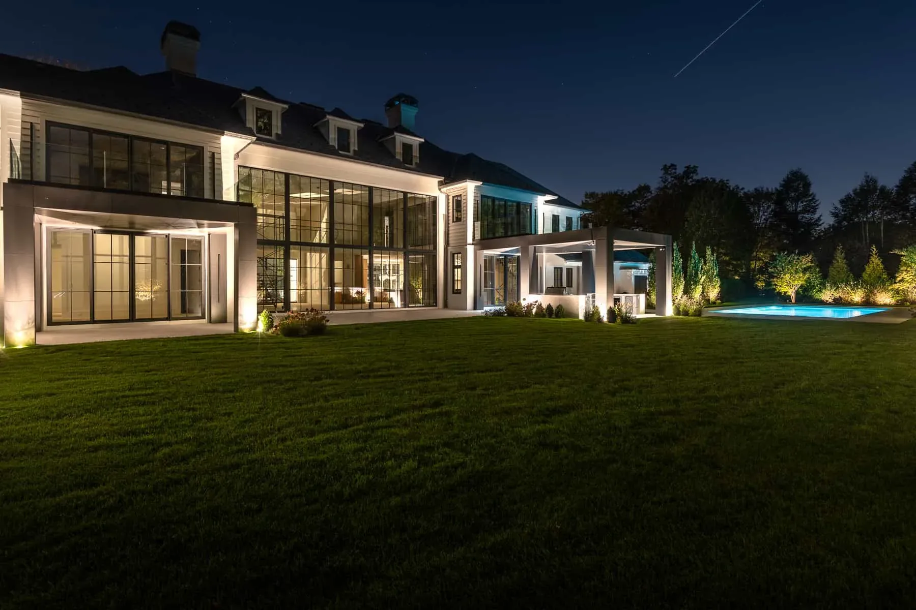 Lit glass-walled rooms opening onto a lawn beside a swimming pool.