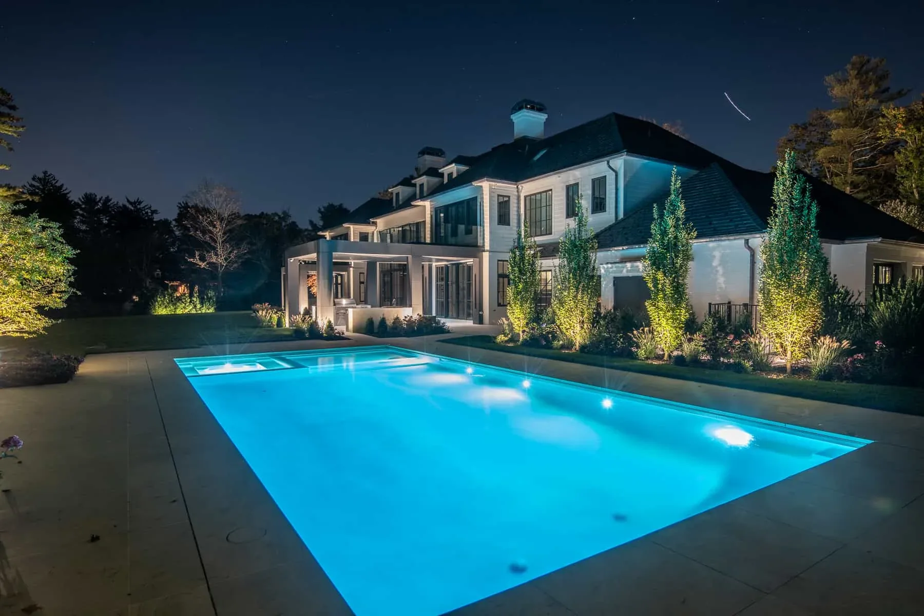 Blue-lit swimming pool beside an illuminated house and garden at night.