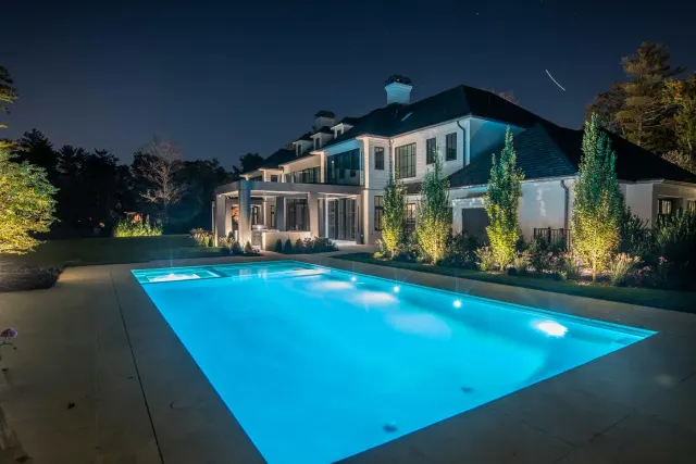 Blue-lit swimming pool beside an illuminated house and garden at night.