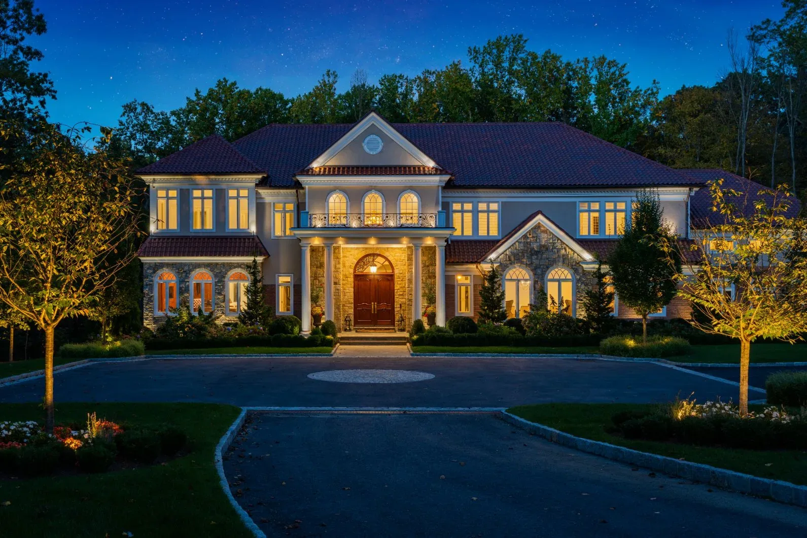 Illuminated stone house and trees facing a curved driveway at night.