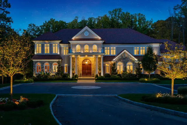 Illuminated stone house and trees facing a curved driveway at night.