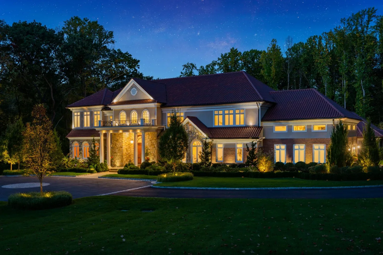 Warm lighting across a large stone house viewed over a lawn at dusk.