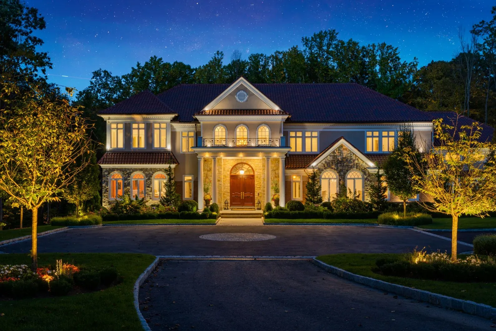 Illuminated stone house and trees facing a curved driveway at night.