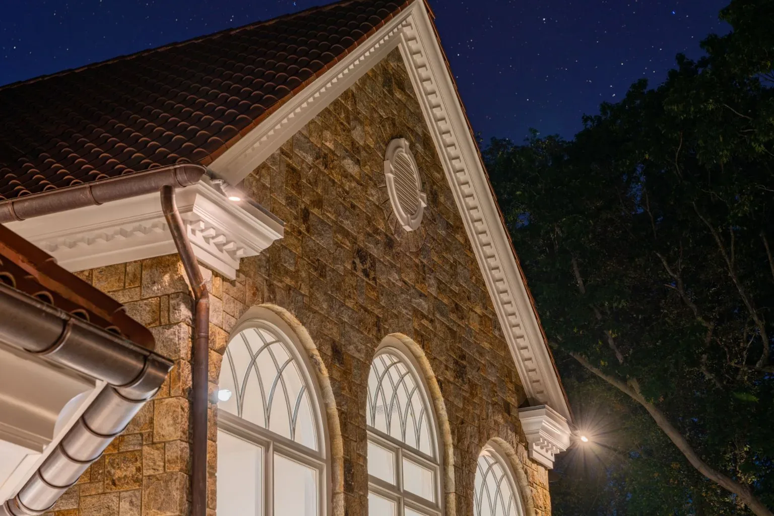 Lighting on a stone gable above two arched windows at night.