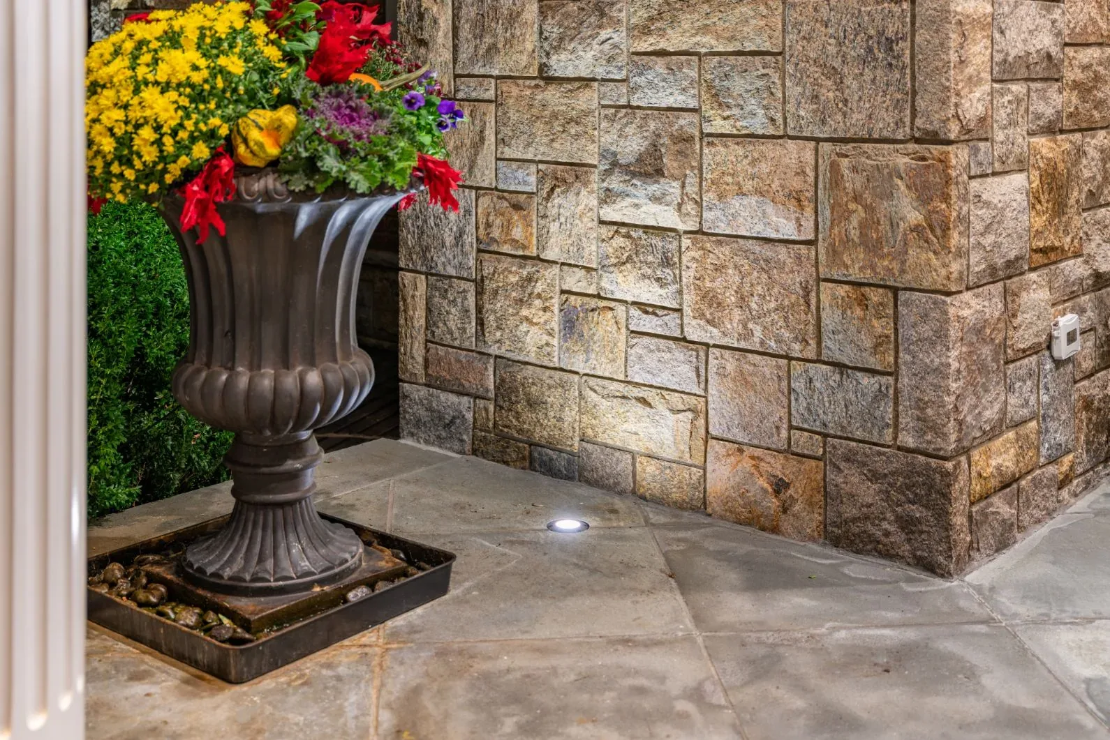 Small ground light illuminating a stone wall beside a large flower planter.