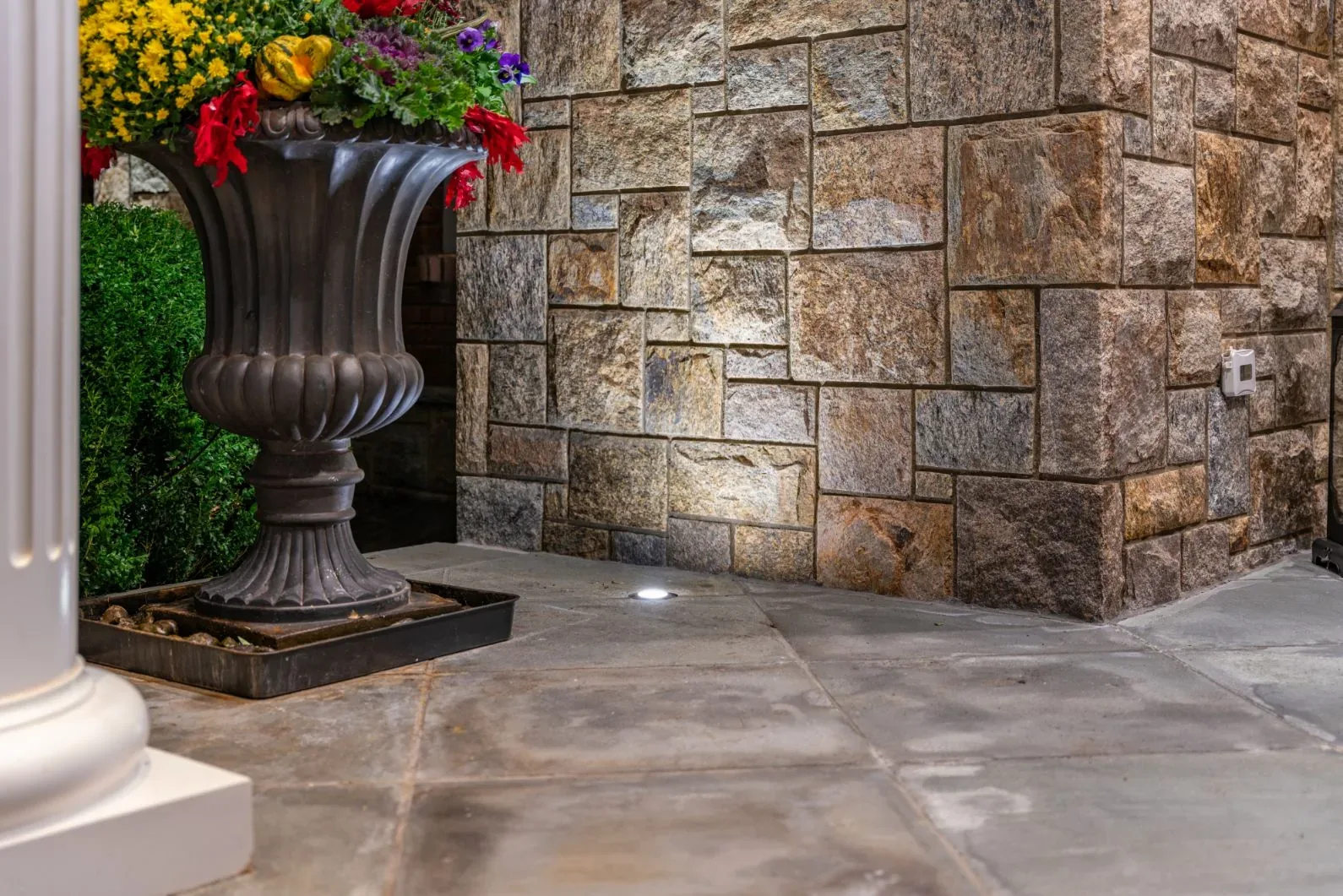 Small ground light illuminating a stone wall beside a large flower planter.
