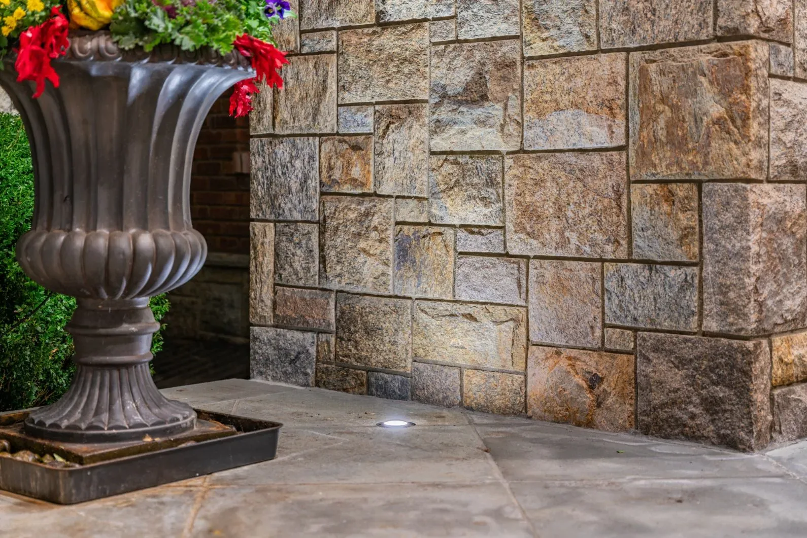 Small ground light illuminating a stone wall beside a large flower planter.
