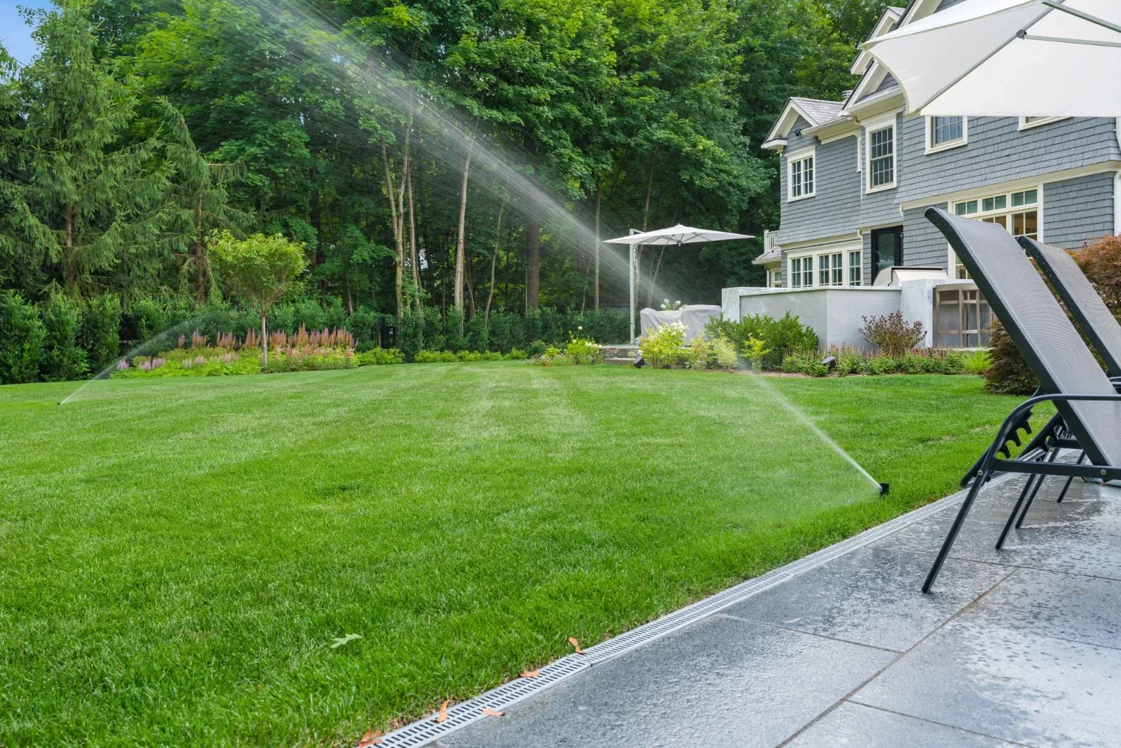 Sprinklers watering a backyard lawn beside a house and planted border.