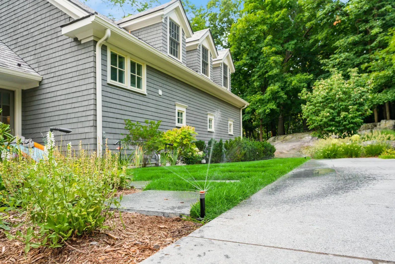 Pop-up sprinkler watering a side lawn and garden beside a house.