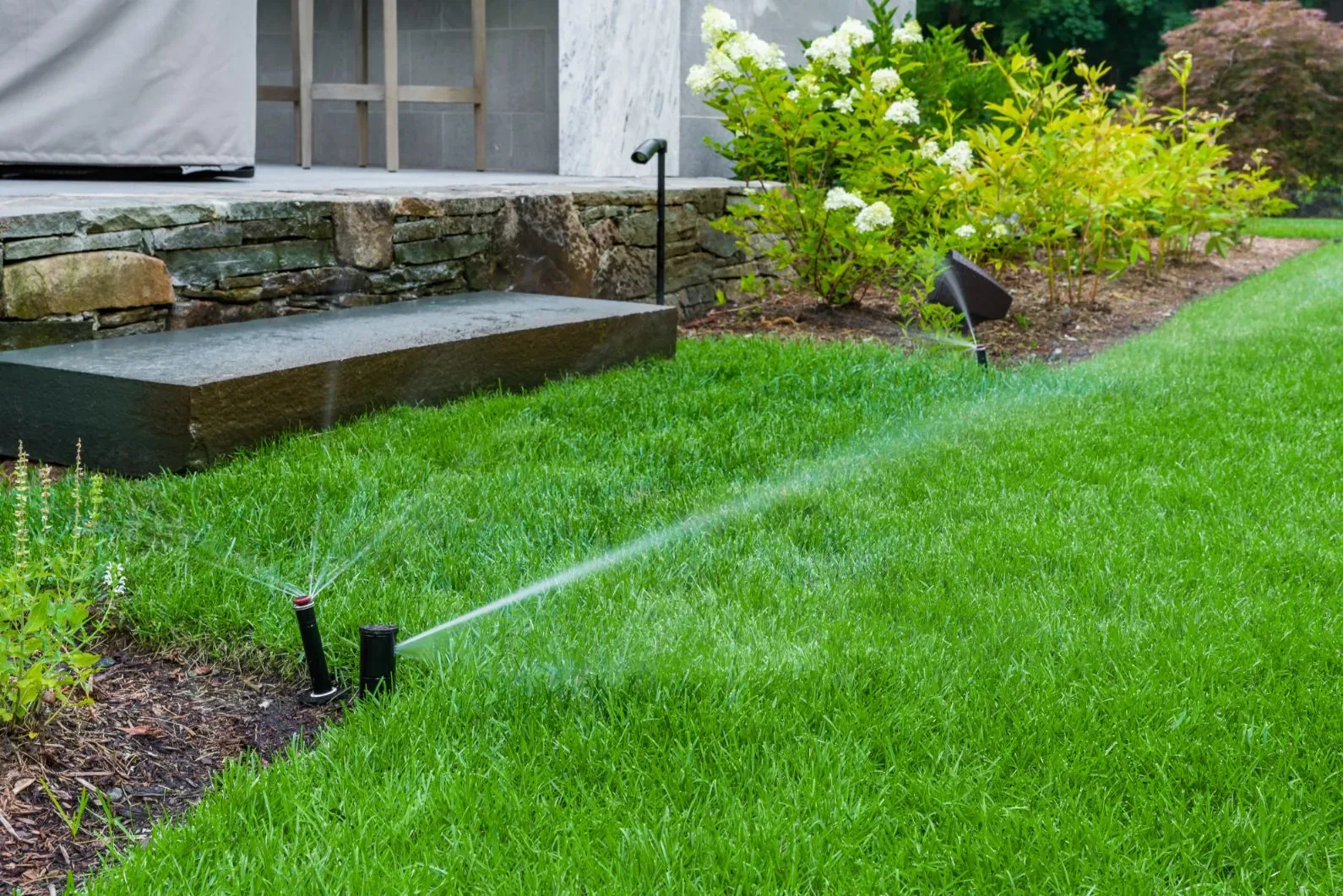 Pop-up sprinkler spraying a lawn beside a patio and garden plants.
