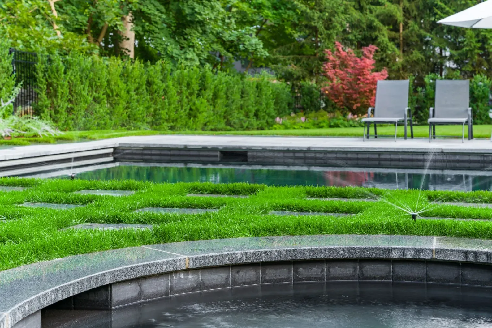 Sprinkler watering grass between a circular spa and swimming pool.