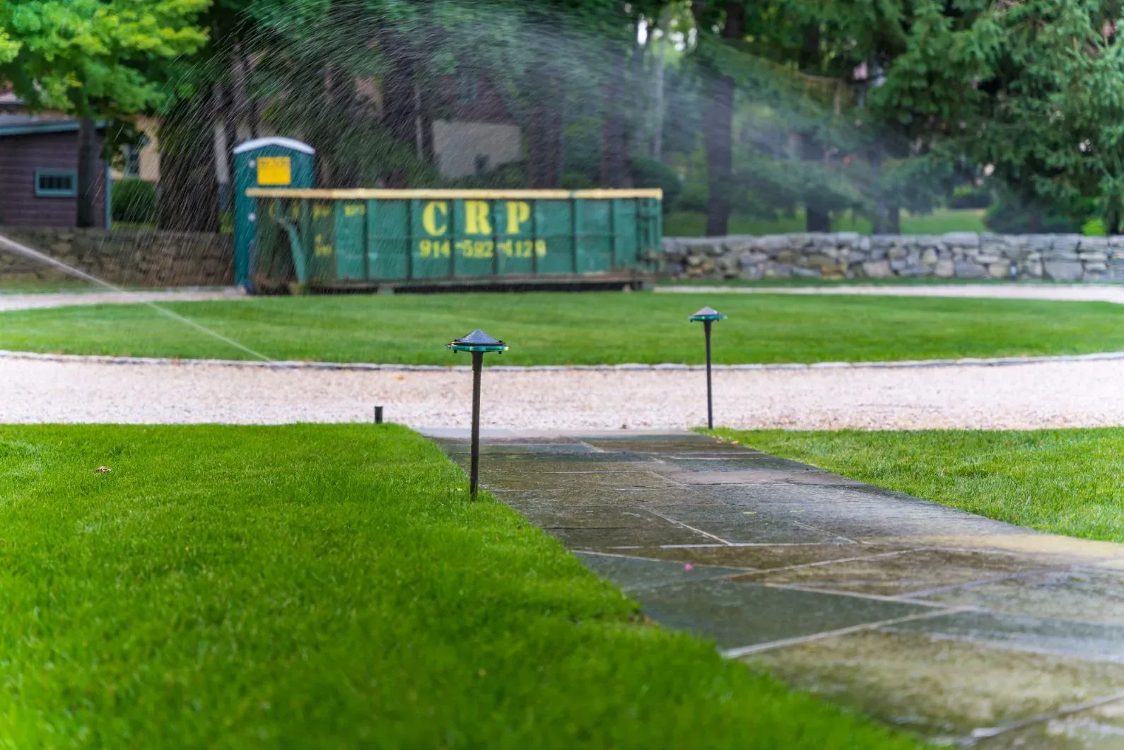 Sprinklers watering a lawn beside a driveway with a dumpster in the background.