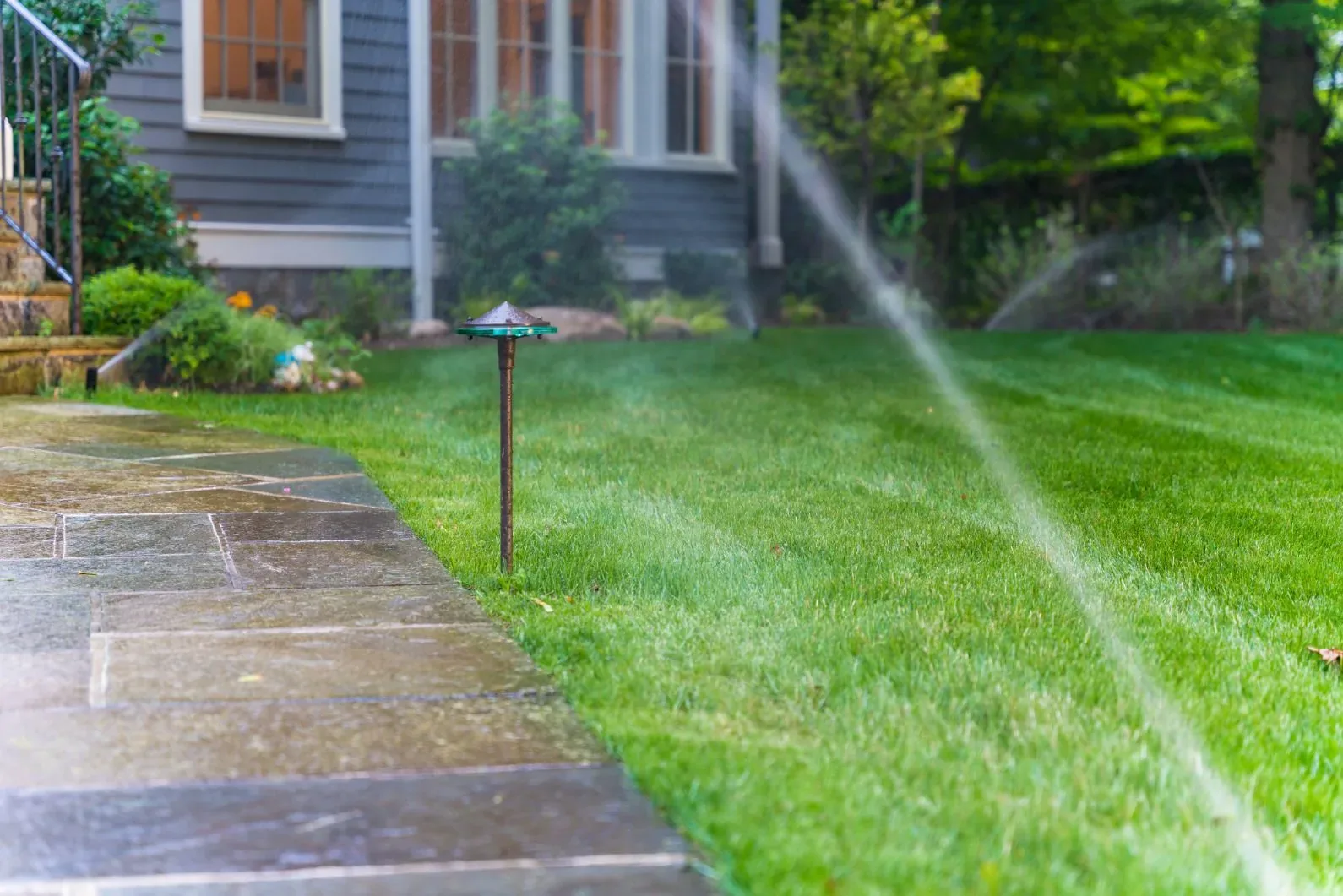 Sprinkler spray across a lawn beside a stone walkway and path light.
