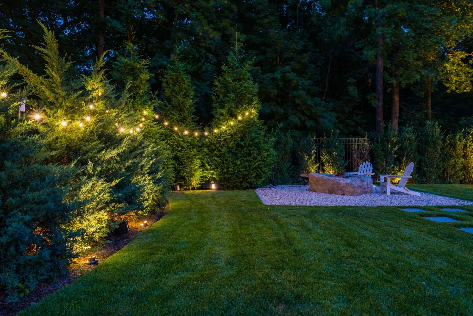 String lights and garden lighting around a lawn and fire pit seating area.
