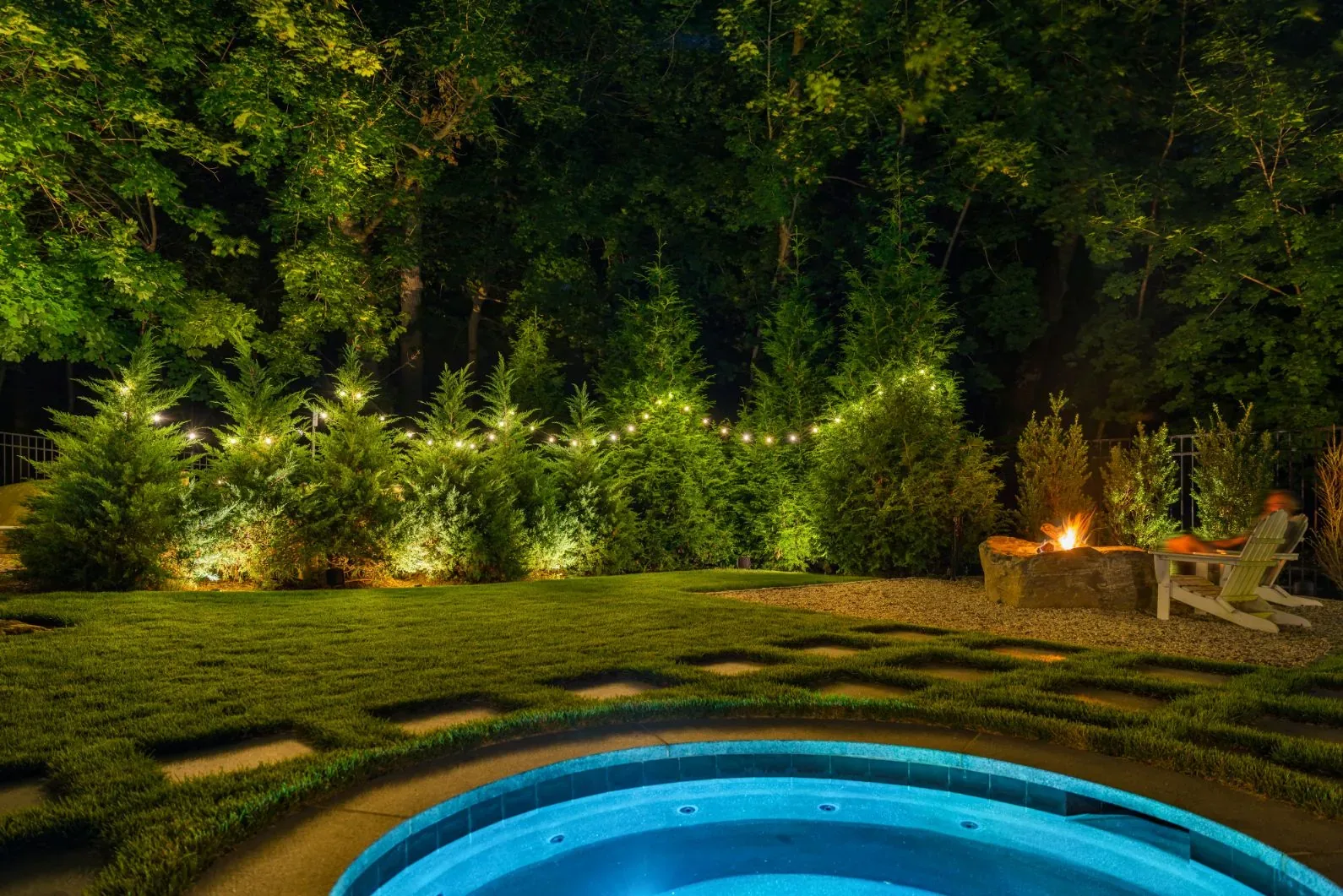 Blue-lit spa beside a stepping-stone lawn and glowing fire pit at night.
