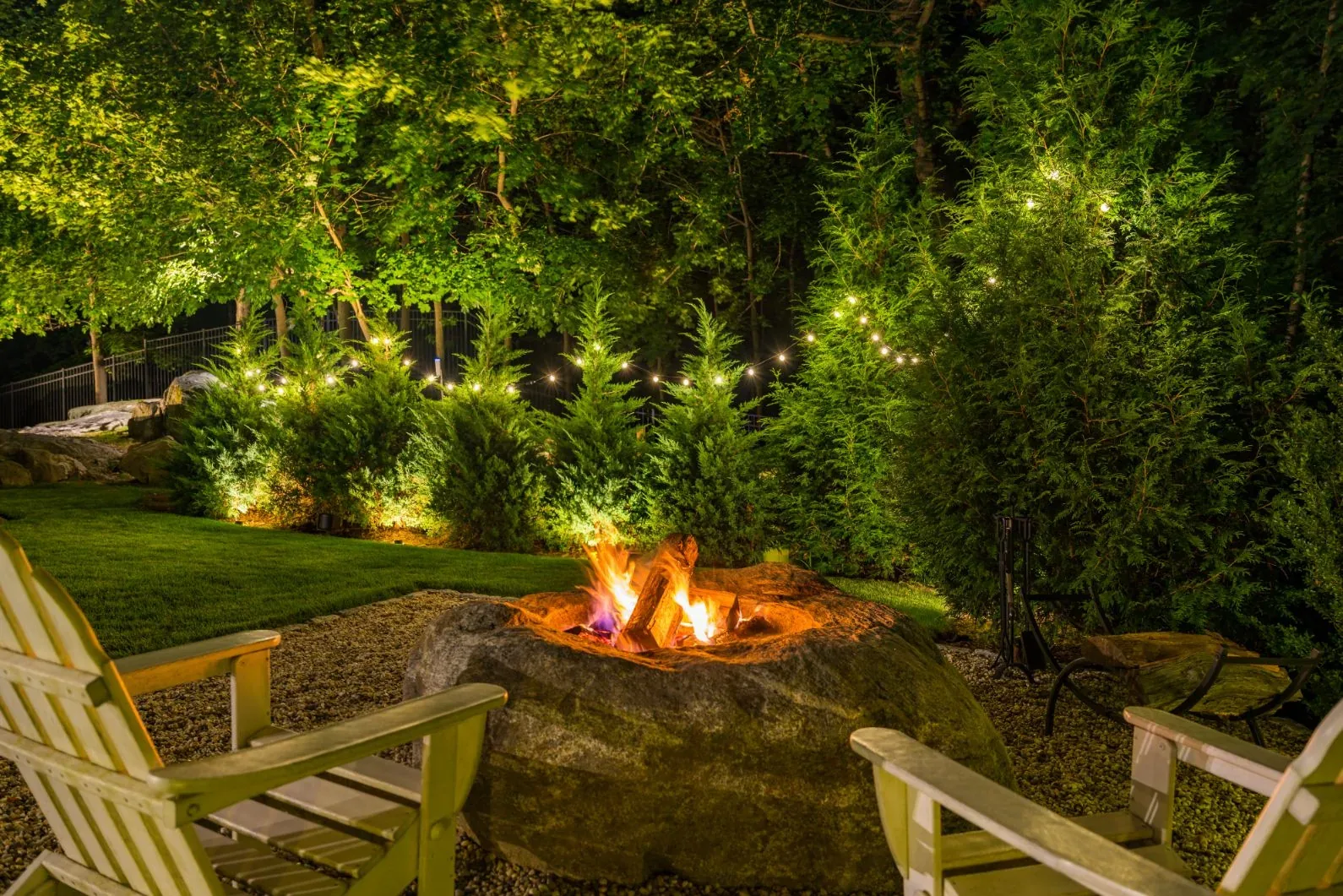 Burning fire pit and chairs in front of illuminated garden shrubs.