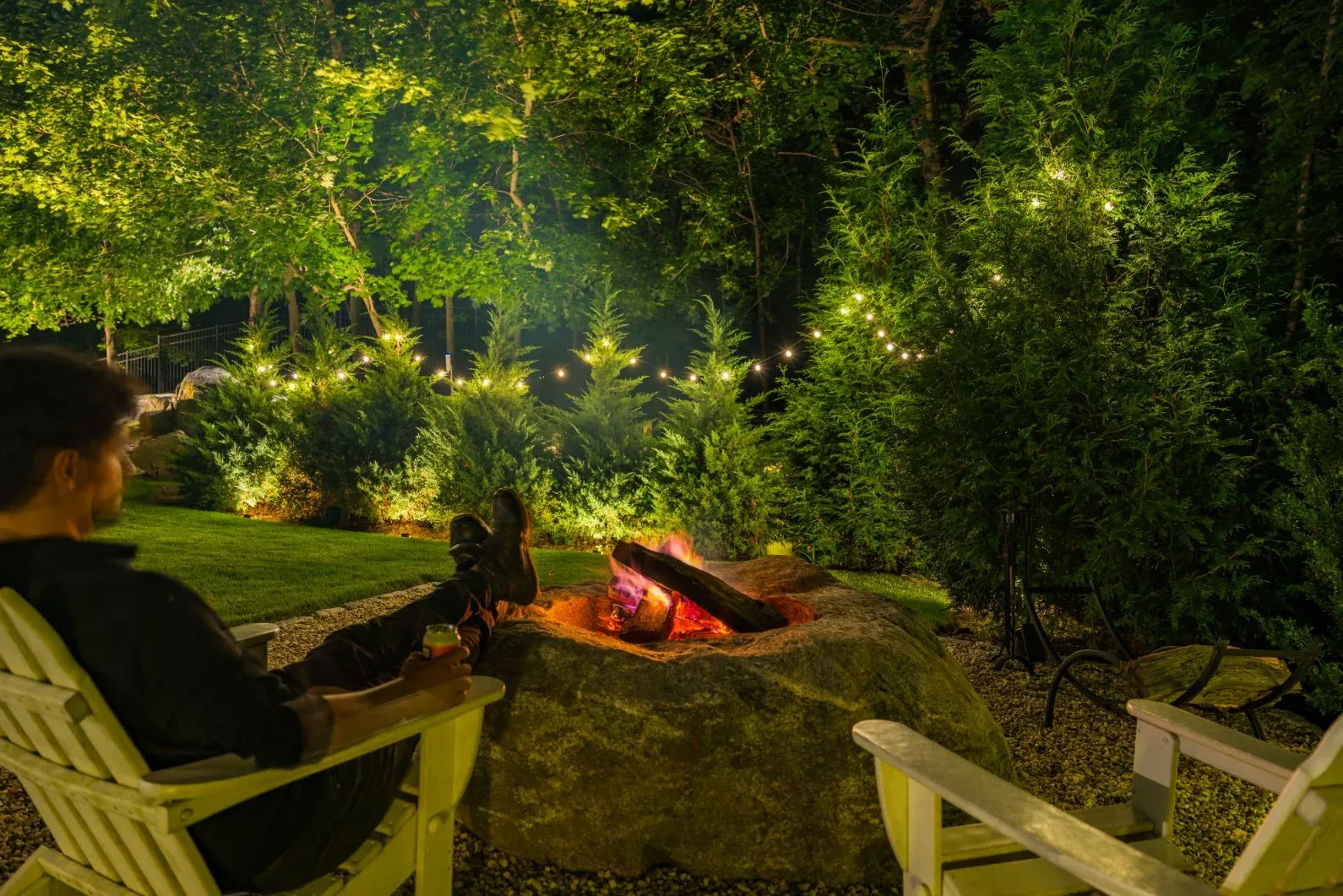 Person seated beside a burning fire pit in an illuminated garden.