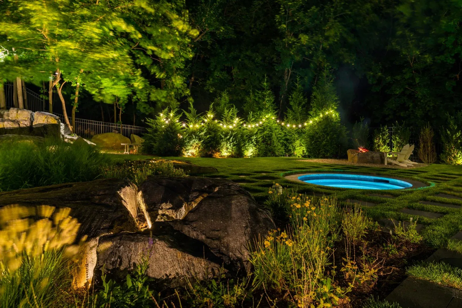 Illuminated rock garden and swimming pool with trees and string lights behind.