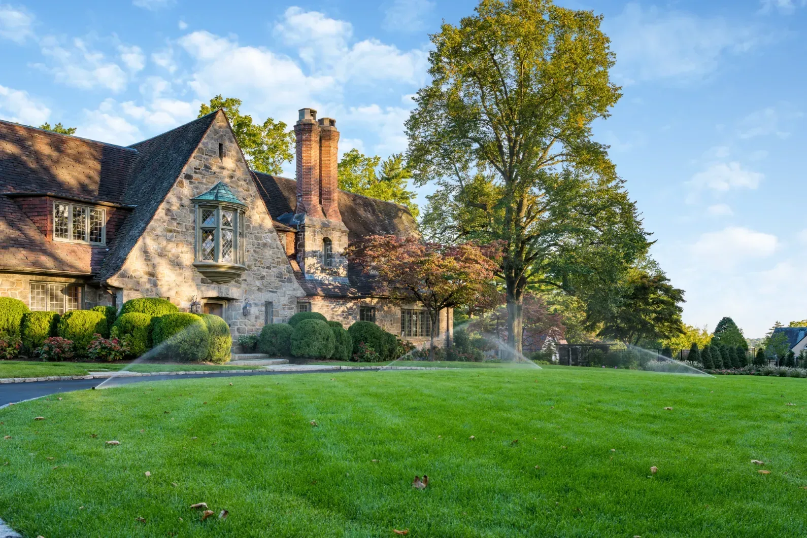 Sprinklers watering a broad lawn in front of a stone house.