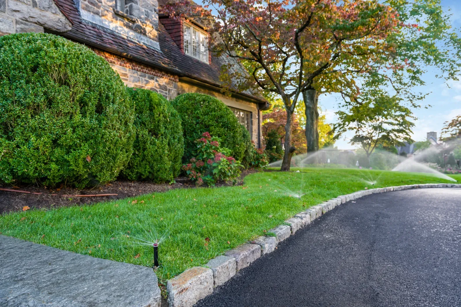 Pop-up sprinklers spraying the lawn beside a curved driveway and stone house.